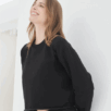 Een vrouw met lichtbruin haar glimlacht tegen een witte muur, gekleed in de Cropped Sweater Ndigo en bijpassende broek.