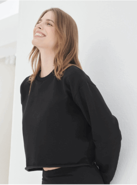 Cropped sweater Adult Een vrouw met lichtbruin haar glimlacht tegen een witte muur, gekleed in de Cropped Sweater Ndigo en bijpassende broek.