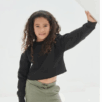 Jong meisje met krullend haar draagt de Cropped Sweater Ndigo en een groene broek, en poseert met één arm opgetrokken tegen een wit oppervlak.