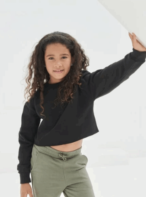 Screenshot 2025-06-05 at 09.52.03 Jong meisje met krullend haar draagt de Cropped Sweater Ndigo en een groene broek, en poseert met één arm opgetrokken tegen een wit oppervlak.