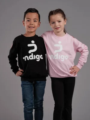 Twee jonge dansers in bijpassende Ndigo Sweatertops poseren tegen een Twee kinderen staan te glimlachen, elk met een Sweater Ndigo aan. De ene is gestyled in een zwarte versie, terwijl de andere gekleed is in roze. Ze poseren tegen een effen grijze achtergrond.