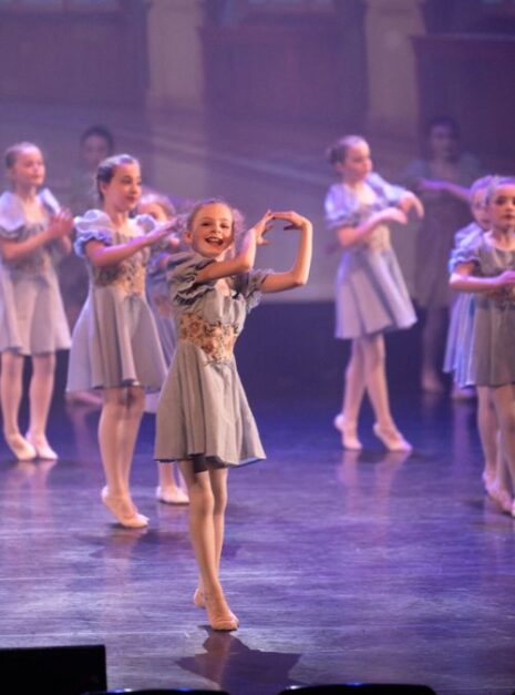 Magic is ND Air 2024 3 Jonge dansers in identieke blauwe kostuums voeren op het podium een balletroutine uit, waarbij één danser op de voorgrond een pose aanneemt. Hun sierlijke bewegingen creëren een magische sfeer.