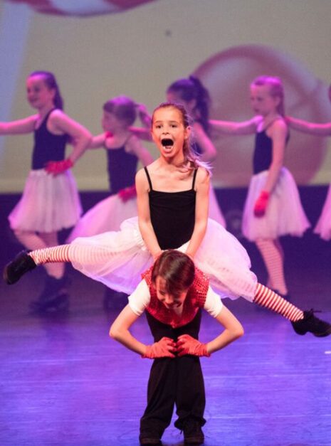 Magic is ND Air 2024 4 Twee jonge dansers treden op het podium op; de een balanceert terwijl hij de splits op de rug van de ander doet. Beiden dragen podiumkostuums met gestreepte sokken en handschoenen, die de magie van ND Air 2024 belichamen. Andere dansers sieren de achtergrond en dragen bij aan de betoverende scène.