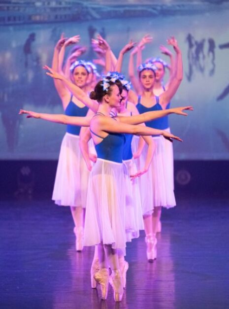 Magic is ND Air 2024 7 Balletdansers in blauwe topjes en witte rokken treden op het podium op, elk met een bloemenhoofddeksel. Tegen de achtergrond van een sereen landschap bewegen ze met moeiteloze gratie alsof er magie in de lucht hangt.