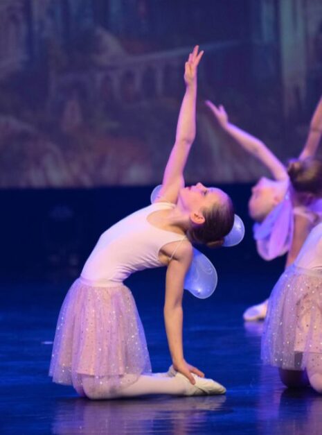 Magic is ND Air 2024 8 Een jonge danseres in een wit kostuum voert een balletpose uit op het podium. Haar gratie legt de magie van ND Air 2024 vast, met één arm omhoog gestrekt.
