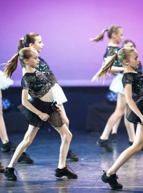 15. FUNKY KIDS 1+2+3 - N-SPIRATION - NDIGO-18 Een groep jonge Ndigo Funky Dance meisjes in glinsterende topjes en rokjes voert een dansroutine uit op het podium tegen een paarse achtergrond.