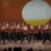 Een grote groep danst op het podium in rode shirts en zwarte broeken tegen een zonnige achtergrond in Video: N-SPIRATION Dansvoorstelling 2025.